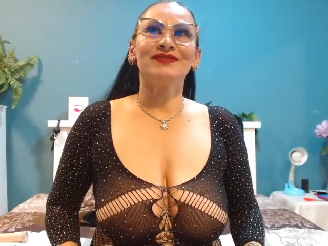 DonnaJones with dark hair, red lipstick, and glasses, wearing a black, see-through, polka-dot dress with a lace-up front, showing her big, round tits and a gold necklace.