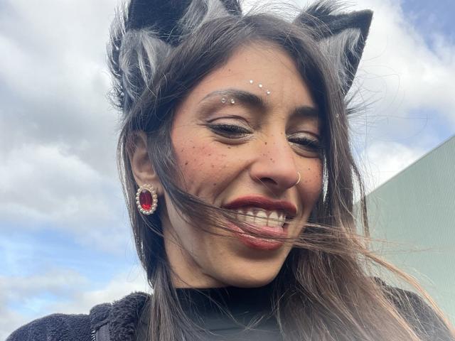 AmanitheWolfy, avec des oreilles de chat, des cheveux foncés et du rouge à lèvres, sourit les yeux fermés, portant des boucles d'oreilles rouges et un pull noir à col roulé. Elle a de petites taches de rousseur et des piercings au nez..