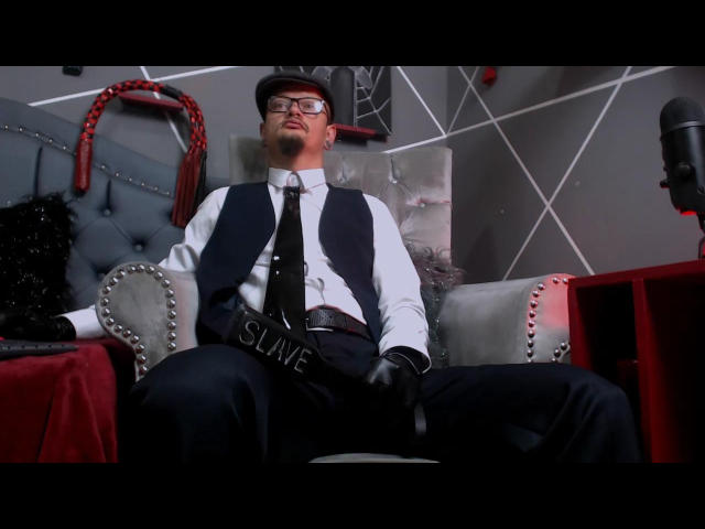 ZainKreiger, wearing glasses and a cap, sits with a fat cock in a black vest, white shirt, and tie, slapping his thick, dark-skinned, erect ass.