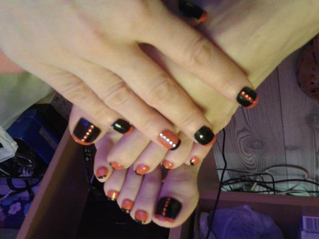 Die Hände von HotSensationX, mit schwarzem Nagellack und orange-weißen, LED-ähnlichen Designs, liegen auf ihren Füßen, die ebenfalls eine ähnliche orange-schwarze Nagelkunst aufweisen..