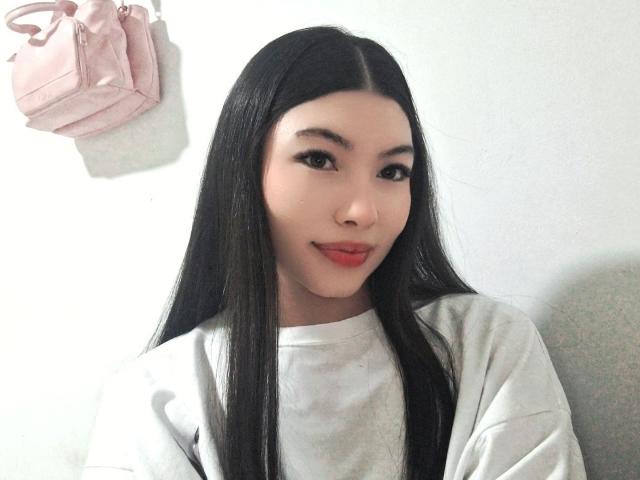 ViolettaLeroy with long black hair, pale skin, and red lipstick, wearing a white shirt, smiling slightly, pink bag hanging on the wall behind her.