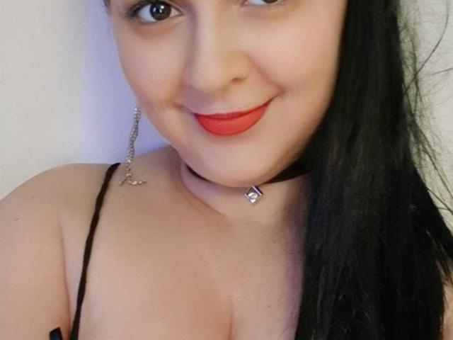 LoraHazel with black hair, red lipstick, and black choker, smiling at camera, showing cleavage in black strap top, small diamond earring, and a slight tan.
