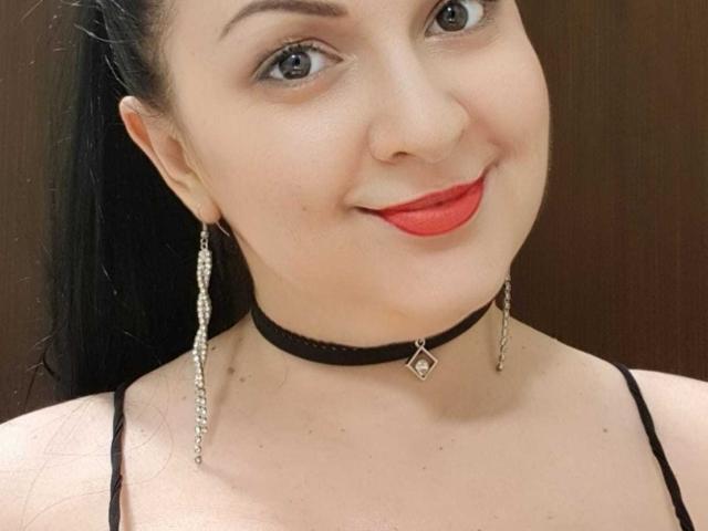 LoraHazel has fair skin, red lipstick, and black hair in a ponytail. She's wearing a black choker with a diamond, and long silver earrings. Her tits are visible.
