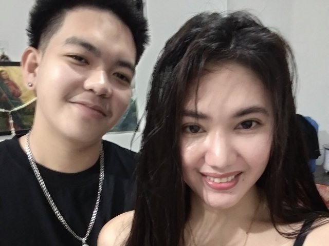 AsianCoupleDesire : un homme asiatique un peu corpulent et une jeune femme asiatique mignonne, tous les deux souriants. Il porte un t-shirt noir et une chaîne, elle a de longs cheveux noirs et un joli visage..
