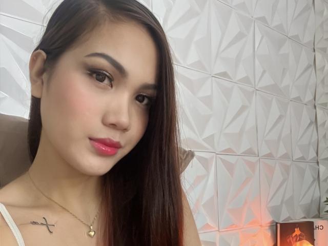 RochelleDee has long brown hair, pink lipstick, and a small tattoo on her left collarbone. She's wearing a white top and a gold necklace.