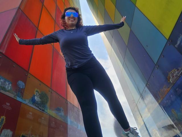 AURORAHOFFMAN with curly hair, blue sunglasses, black long-sleeve shirt, black pants, and white sneakers, standing with arms out, smiling, against colorful metal panels.