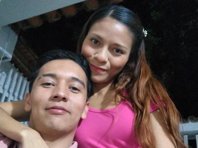 KarolAndLenny with long brown hair, pink top, and small tits, standing behind KarolAndLenny with short black hair, smiling. Both have light skin and are in a night photo.