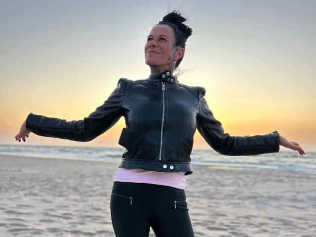 LucyLove-hot stands on a beach, arms out, wearing a black leather jacket and black pants, with a pink shirt underneath, smiling at a sunset.