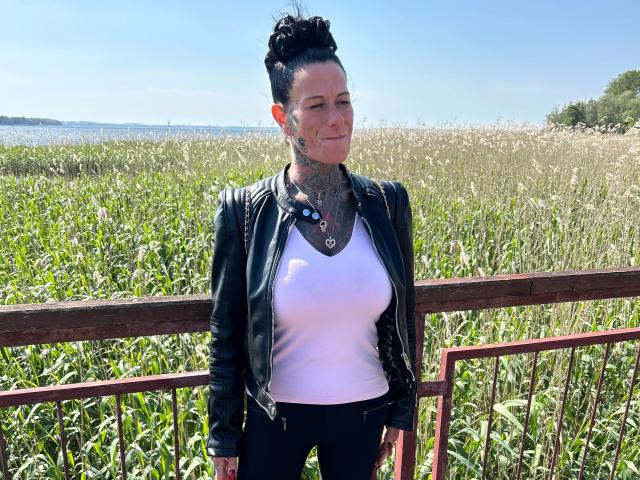 LucyLove-hot stands with a black leather jacket, white shirt, and black pants, showing her tits and tattoos, in a grassy field with a wooden fence in the background.