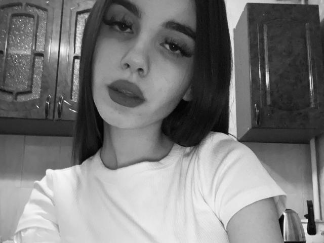 Selfie in bianco e nero di KathleenLois con lunghi capelli scuri, sopracciglia folte, eyeliner e rossetto scuro, che indossa una maglietta bianca, mostrando il suo viso e la parte superiore del petto..
