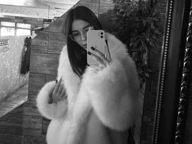 KathleenLois mit langen, dunklen Haaren, Brille und einem flauschigen weißen Mantel, die ein Selfie mit ihrem Handy macht, wobei schwarze Nägel und ein leichtes Lächeln zu sehen sind. Brüste sind teilweise bedeckt..
