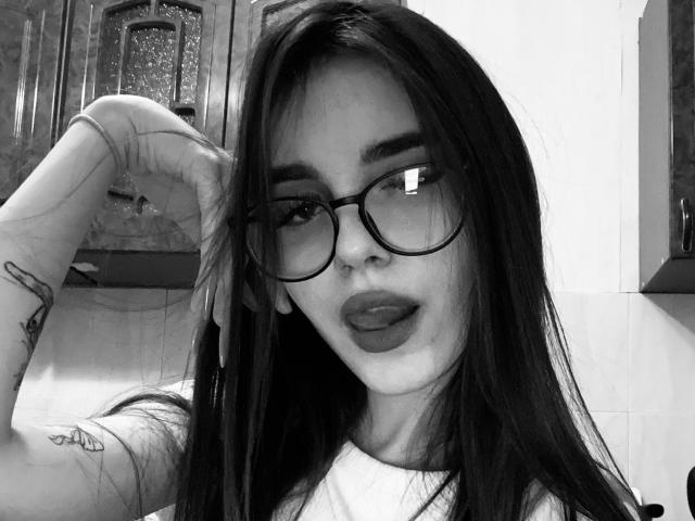 Selfie in bianco e nero di KathleenLois con lunghi capelli scuri, occhiali, piercing al naso, tatuaggi sul braccio e labbra carnose, con una mano tra i capelli, che indossa una camicia bianca..