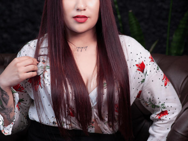 ValeriaTorrez met lang rood haar, rode lippenstift, een witte blouse met bloemenprint, een tatoeage op de borst, een getatoeëerde arm, die een blouse vasthoudt, een bruine leren bank, kijkt naar beneden, zacht licht, sexy, aantrekkelijk..