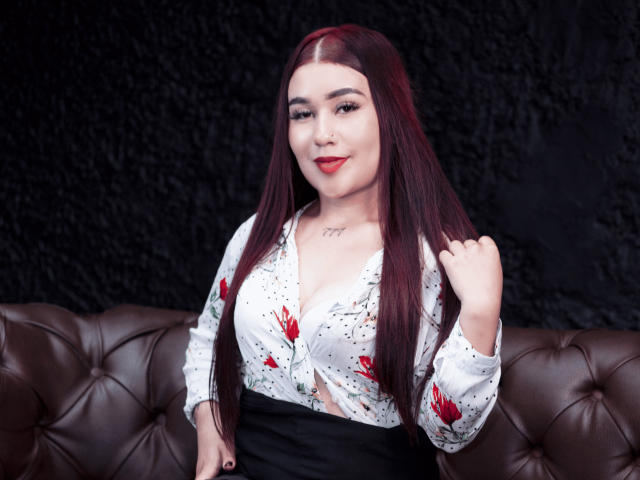 ValeriaTorrez with long red hair, white floral blouse, black skirt, red lipstick, holding hair, showing small tits, sitting on brown leather couch, black textured background.
