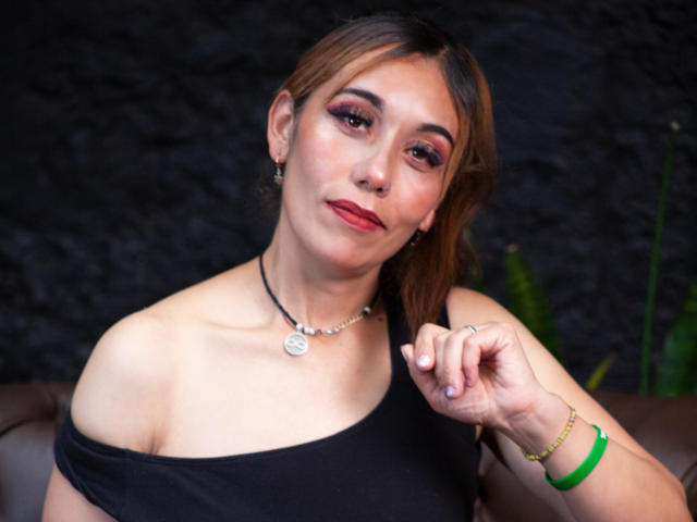 MadeleineSartori with light brown hair, red lipstick, and dark makeup, wearing a black off-shoulder top, necklace, green bracelet, and earrings, slightly raising her right hand.