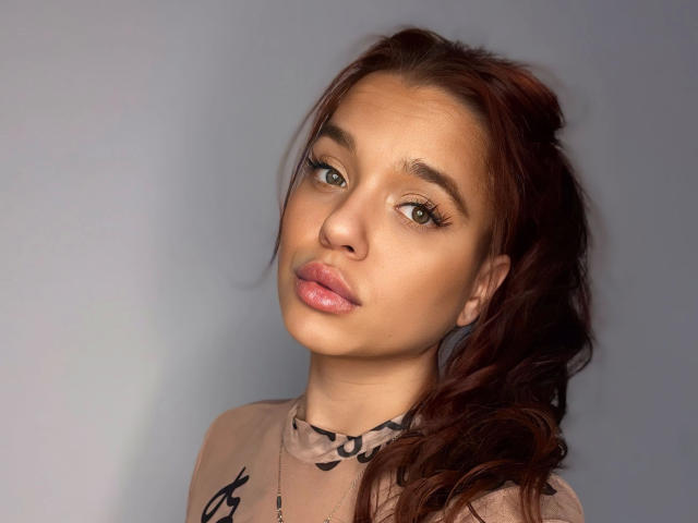 Babilonian-hot with pouty pink lips, brown eyes, and wavy red hair in a ponytail, wearing a sheer black top, has a cute, sexy face.