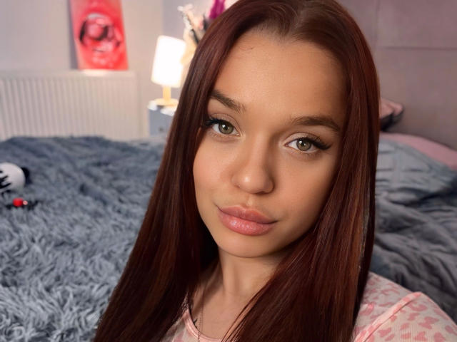 Babilonian-hot with straight brown hair, green eyes, and pink lips, wearing a pink top, has a cute face and a slight smile in a bedroom with a blue bed.