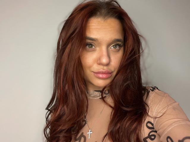 Babilonian-hot with long, wavy auburn hair, light brown eyes, and pink lips, wearing a sheer, pink top with black patterns, and a cross necklace, looking at the camera.
