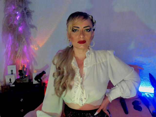 JeaninexScott with blonde ponytail, white ruffled blouse, red lipstick, black lace panties, earrings, and makeup, sitting on bed with dildo and vibrator, sexy and confident.