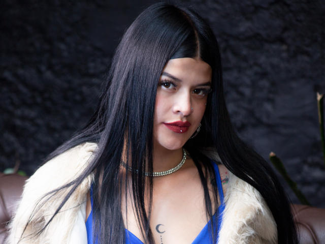 Luciana Dartois, com cabelos longos e pretos, blusa azul, batom vermelho, colar de pérolas, casaco de pele branca, pequena tatuagem em forma de crescente no peito, cadeira de couro marrom, fundo escuro com textura..