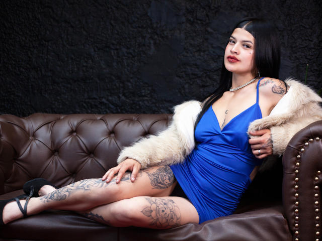 Luciana Dartois, com tatuagens e aparência sensual, está sentada em um sofá de couro marrom, vestindo um vestido azul, um casaco de pele sem mangas e saltos pretos, olhando para a câmera com um sorriso confiante..