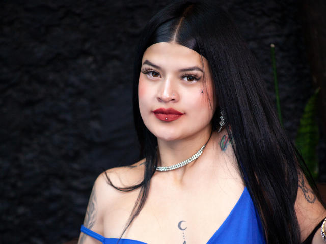 Luciana Dartois mit langem, schwarzem Haar, blauem Kleid, rotem Lippenstift, Nasenpiercing, tätowierter Brust, einer Perlenkette und Ohrringen, lächelnd in die Kamera. Brüste sichtbar, dunkler Hintergrund..
