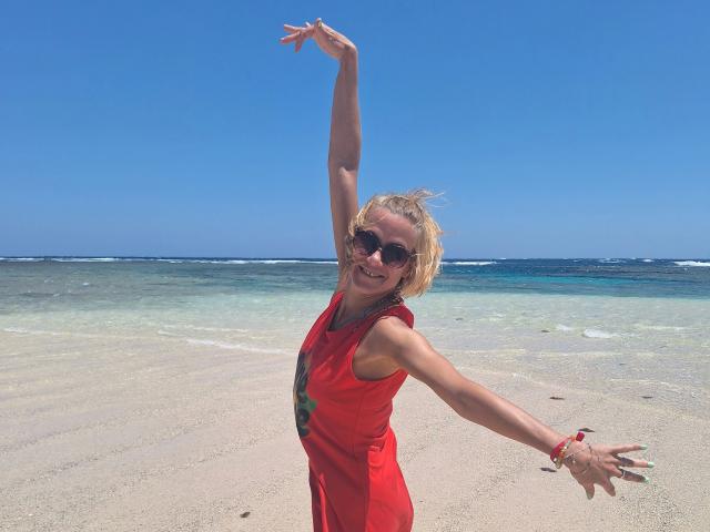 xcYola-hot with blonde hair, wearing a red dress, raising one arm, and smiling at the beach. She's got dark sunglasses and a bracelet on her right wrist.