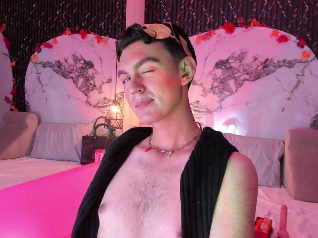 DeusBeauty, shirtless, with black hair and a headband, winking, has a silver necklace, and a red mark on his chest, in a pink-lit bedroom with a map on the wall.