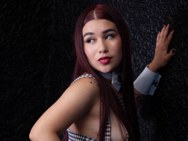 ValeriaTorrez, con i capelli rossi, che si guarda indietro, indossa una camicetta a quadri, mostra una parte del seno, ha un tatuaggio sul braccio, una mano appoggiata su un muro scuro e strutturato, rossetto rosso, smalto nero per unghie, espressione seria..