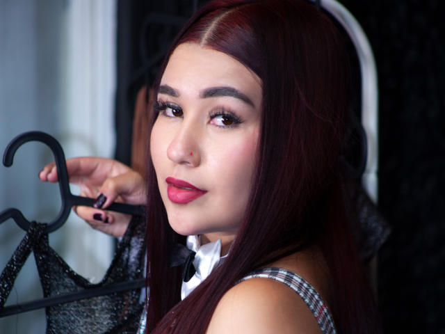 ValeriaTorrez, avec ses longs cheveux roux, son rouge à lèvres rouge et son eye-liner noir, tient un soutien-gorge noir scintillant et regarde par-dessus son épaule avec un léger sourire. Elle a des ongles foncés..