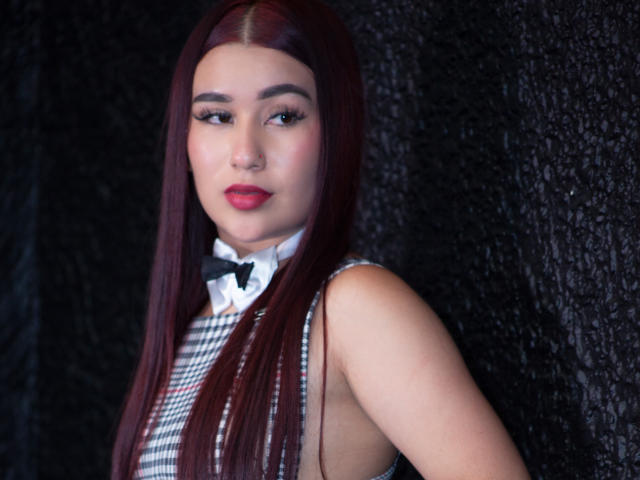 ValeriaTorrez tem cabelos ruivos longos, veste um vestido xadrez com uma gravata borboleta preta e batom vermelho, e está em frente a um fundo escuro com textura, olhando para a câmera..