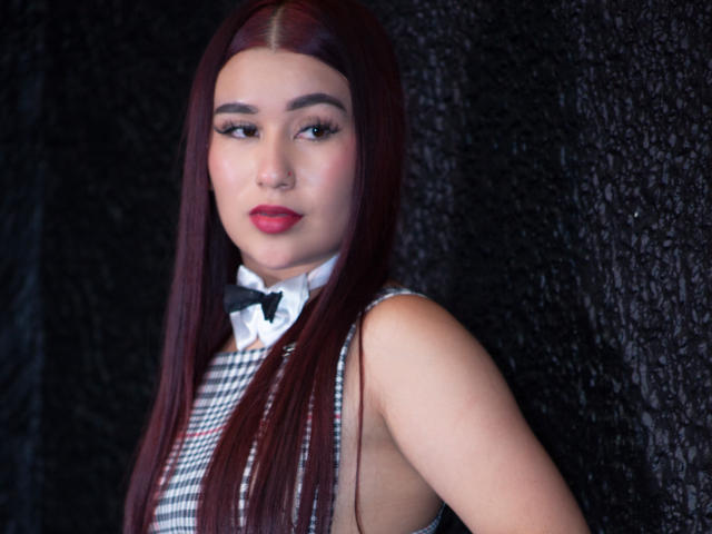 ValeriaTorrez, com cabelos ruivos longos, vestindo uma blusa quadricula preta e branca, um laço preto e batom vermelho, exibindo suas nádegas com as costas levemente viradas..