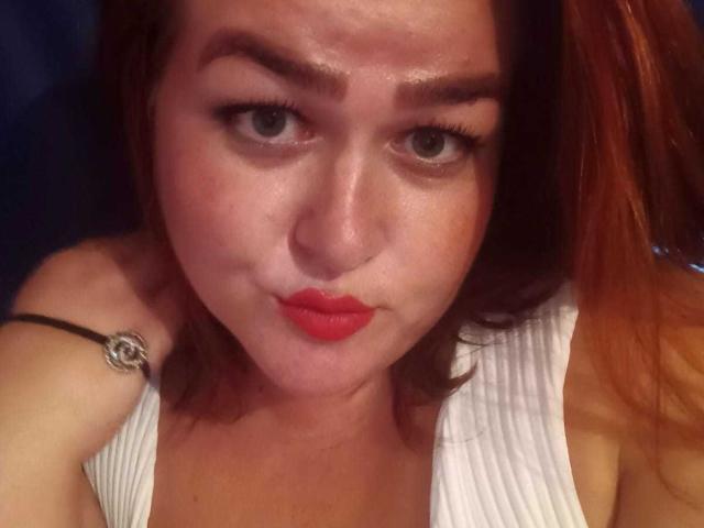 BonnyCarry with red lipstick, slightly puffed cheeks, and raised eyebrows, wearing a white sleeveless top, showing her shoulder with a black bracelet. Her brown hair is partially visible.