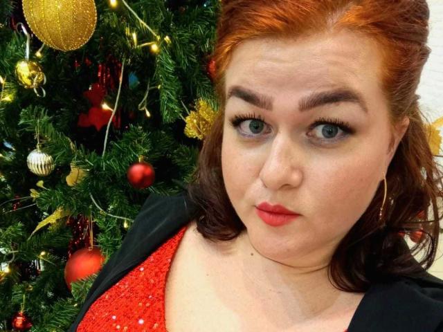 BonnyCarry has red hair, blue eyes, and is wearing red lipstick with a black jacket over a red sequined top. She stands in front of a Christmas tree.