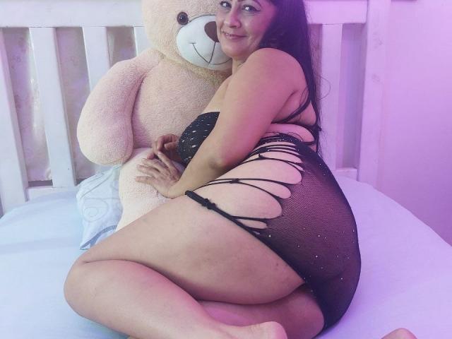 CandelaFoxy, volslank met een groot achterwerk, draagt zwart, versleten kanten lingerie, knielt op een bed en omarmt een grote teddybeer, glimlachend, met lang donker haar, en roze verlichting..