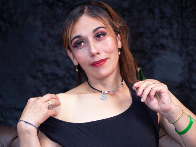 Madeleine Sartori, con i capelli castani, rossetto rosso e trucco, indossa una maglietta nera con una spalla scoperta, tirandola giù per mostrare un seno piccolo, una collana e un braccialetto verde..