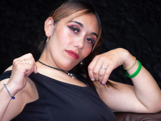 Madeleine Sartori, con i seni leggermente scoperti, che alza la sua maglietta nera, indossa rossetto rosso, un braccialetto verde e diversi anelli, su uno sfondo scuro..