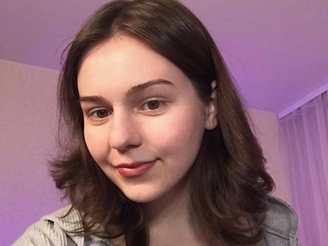 AliceNour has pale skin, brown eyes, and brown wavy hair, with a slight smile showing pink lips. She's wearing a grey shirt, with a subtle purple room light.