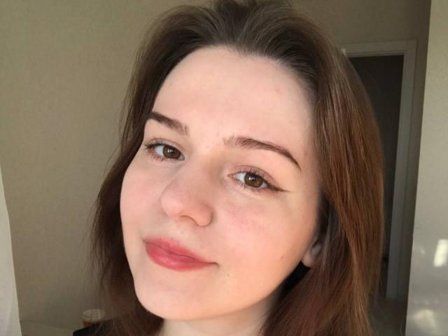 AliceNour has light skin, brown eyes, and brown hair. She's smiling with a small nose and full lips, showing a cute, natural look with a slight blush.