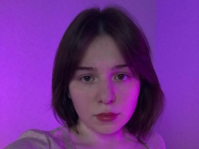 AliceNour with pink-purple lit face, short brown hair, red lips, and subtle makeup, wearing a gray top, looking straight at the camera. Boobless, no visible ass or pussy.