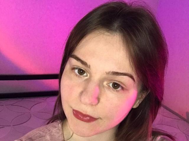 AliceNour with pink and purple-tinged light on her face, brown eyes, dark brown hair, and shiny red lips, wearing a pink shirt, looking at the camera.