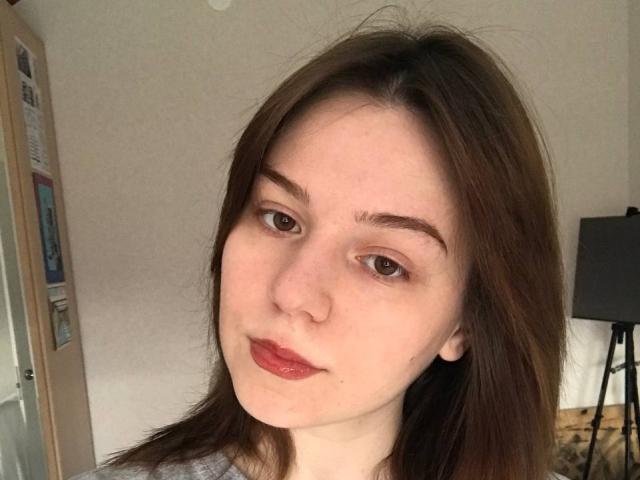 AliceNour has fair skin, brown eyes, and brown hair. She's pouting with red lips and a small beauty mark below her right eye. She's wearing a gray shirt.