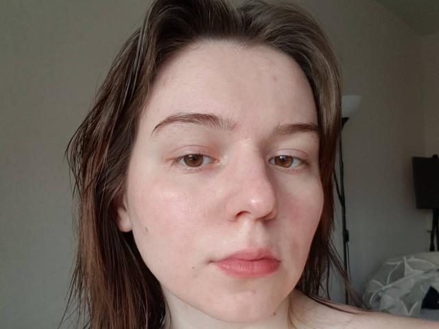 AliceNour has wet brown hair, light skin, brown eyes, and slightly pink lips with a small pimple on her right cheek. She looks straight into the camera.