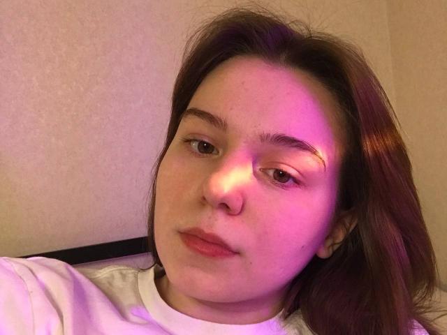 AliceNour has pinkish-purple light on her face, brown eyes, and a nose ring. She's wearing a white shirt, with brown hair and a neutral expression.