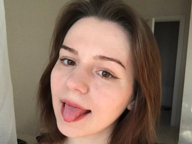 AliceNour's face with brown eyes, fair skin, and brown hair, tongue sticking out, wearing black top, slight freckles, subtle eyeliner, in a well-lit room, looking playful.
