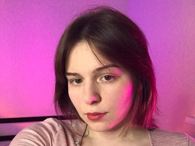 AliceNour has pale skin, brown eyes, and dark brown shoulder-length hair. She's wearing a pinkish shirt with subtle makeup and a red lipstick. Pink and purple light casts on her.