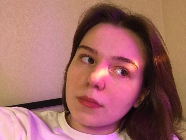AliceNour with pinkish-purple lighting on her face, brown eyes, and slightly parted pink lips, wearing a white shirt, has medium-length brown hair, and a neutral expression.
