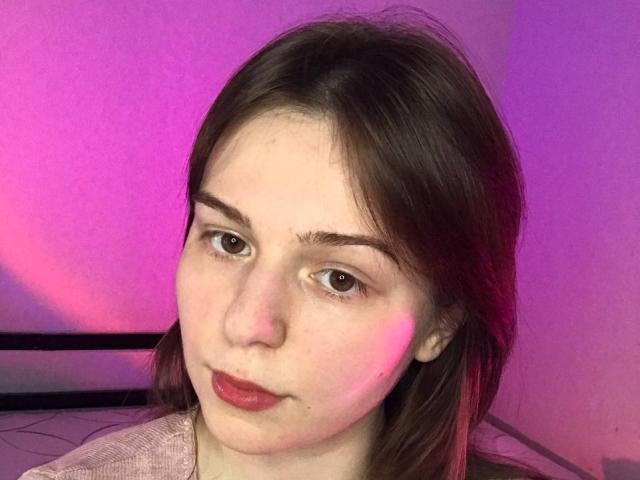 AliceNour has fair skin, brown eyes, and red lipstick with slight pink light on her face. She's wearing a pinkish top, with brown hair covering her ears.