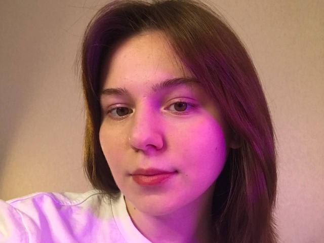 AliceNour's face with pinkish-purple light on right side, brown eyes, slightly parted pink lips, straight brown hair, wearing white shirt, soft facial expression, fair skin, slight freckles.