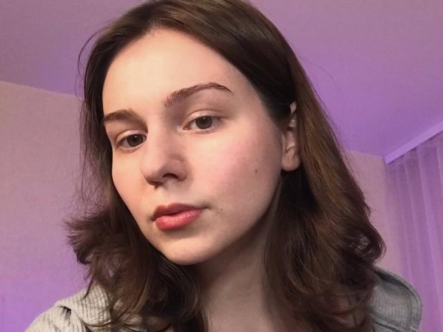 AliceNour with puffy pink lips, brown wavy hair, and thin eyebrows, looking slightly down with a neutral expression, wearing a gray top.